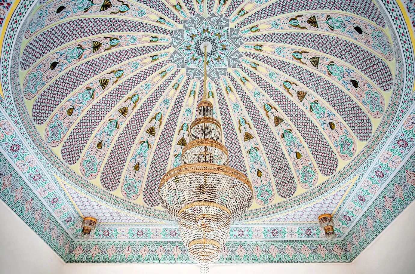 A large golden chandelier against a variegated ceiling adorned with traditional Islamic religious ornamentation.
